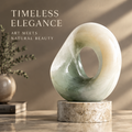Natural jade stone sculpture with open ring form, displayed on a marble-textured stone base in warm ambient light — Timeless Elegance, Art Meets Natural Beauty